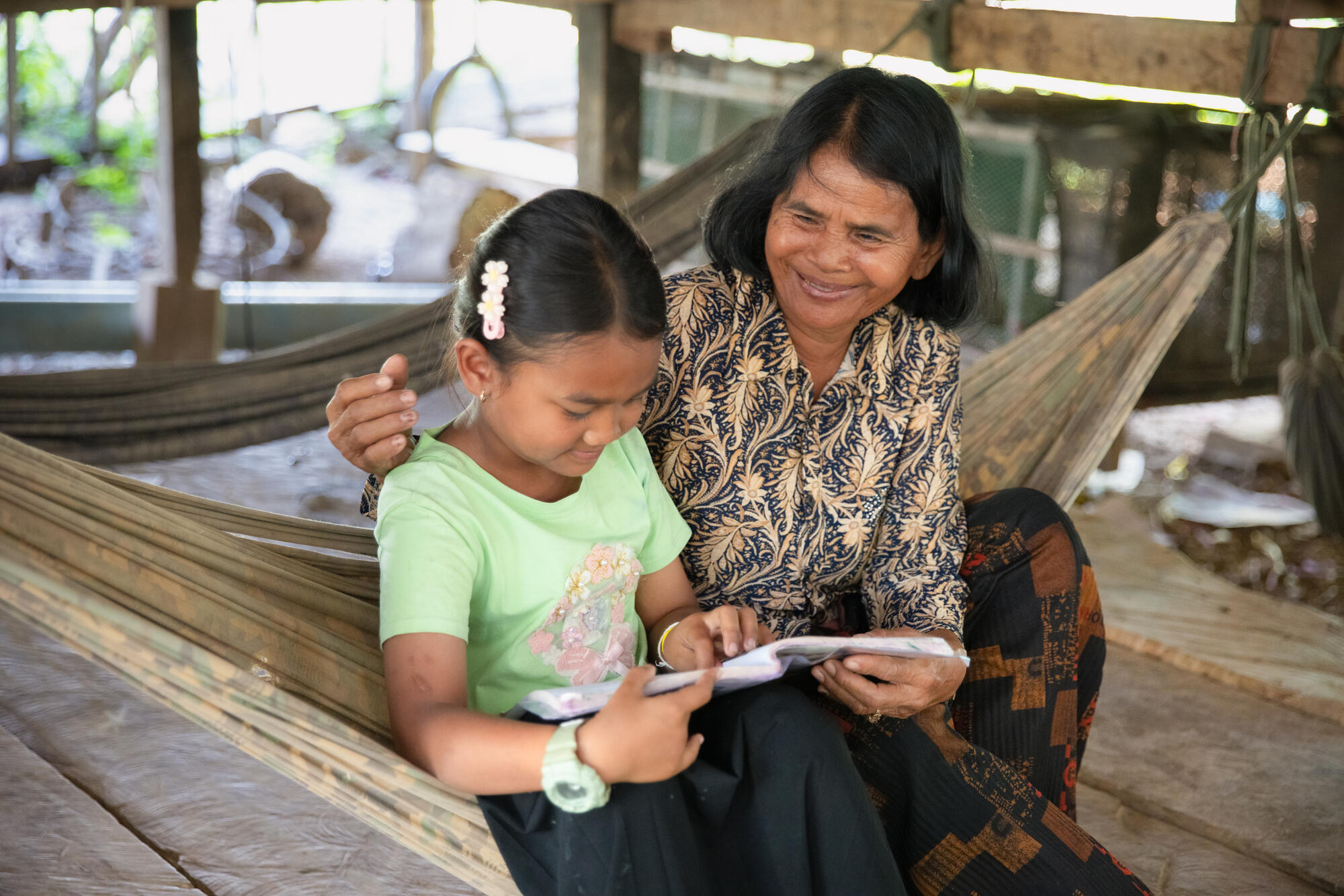 A young girl concentrates on her reading and her grandmother embraces her with pride. They’re both sitting comfortably in a hammock, smiling.