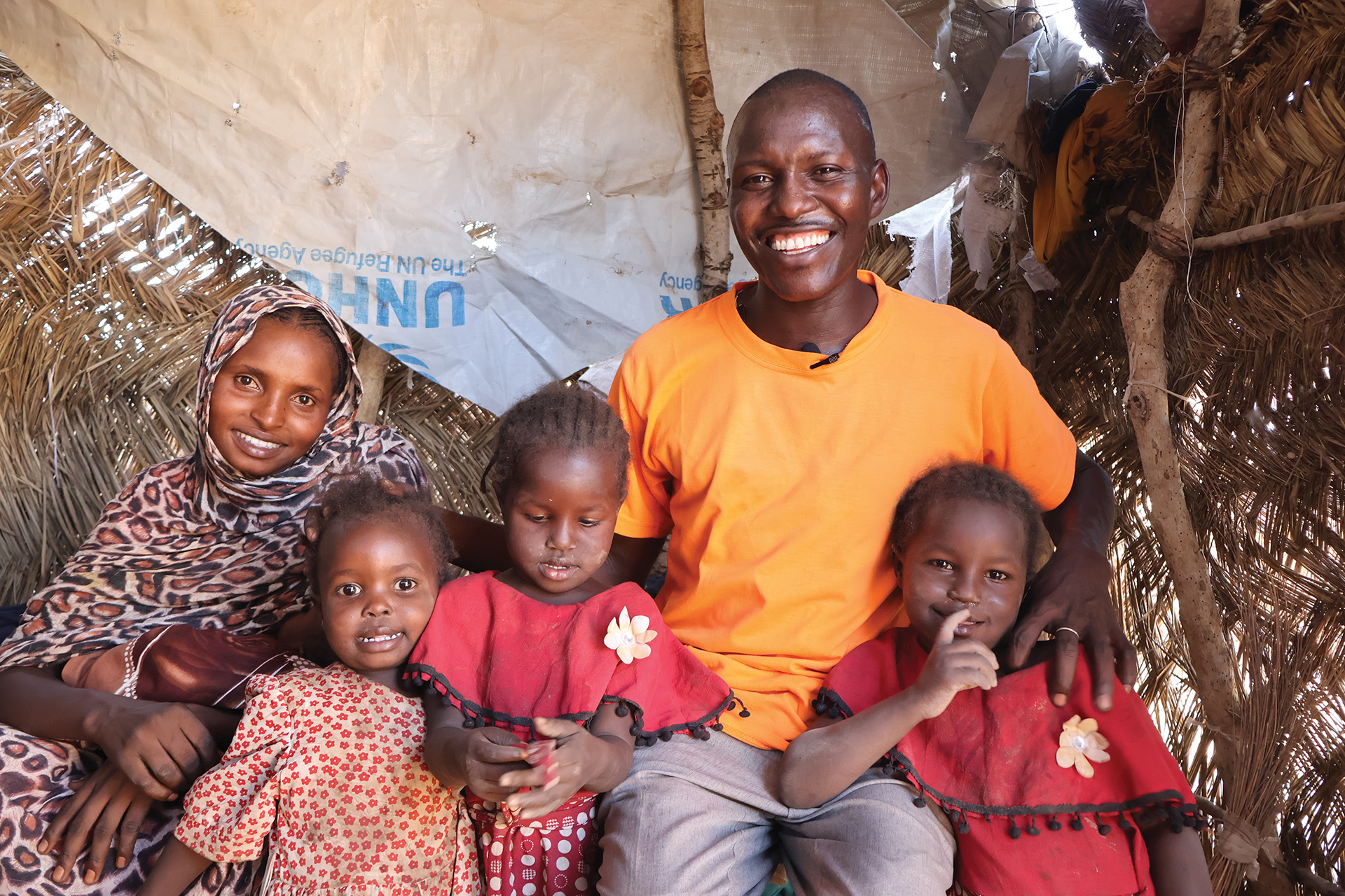 A man sits with three children and his wife, who holds a baby. They’re all smiling to the camera in their modest shelter, a tarpaulin sheet visible in the corner.
