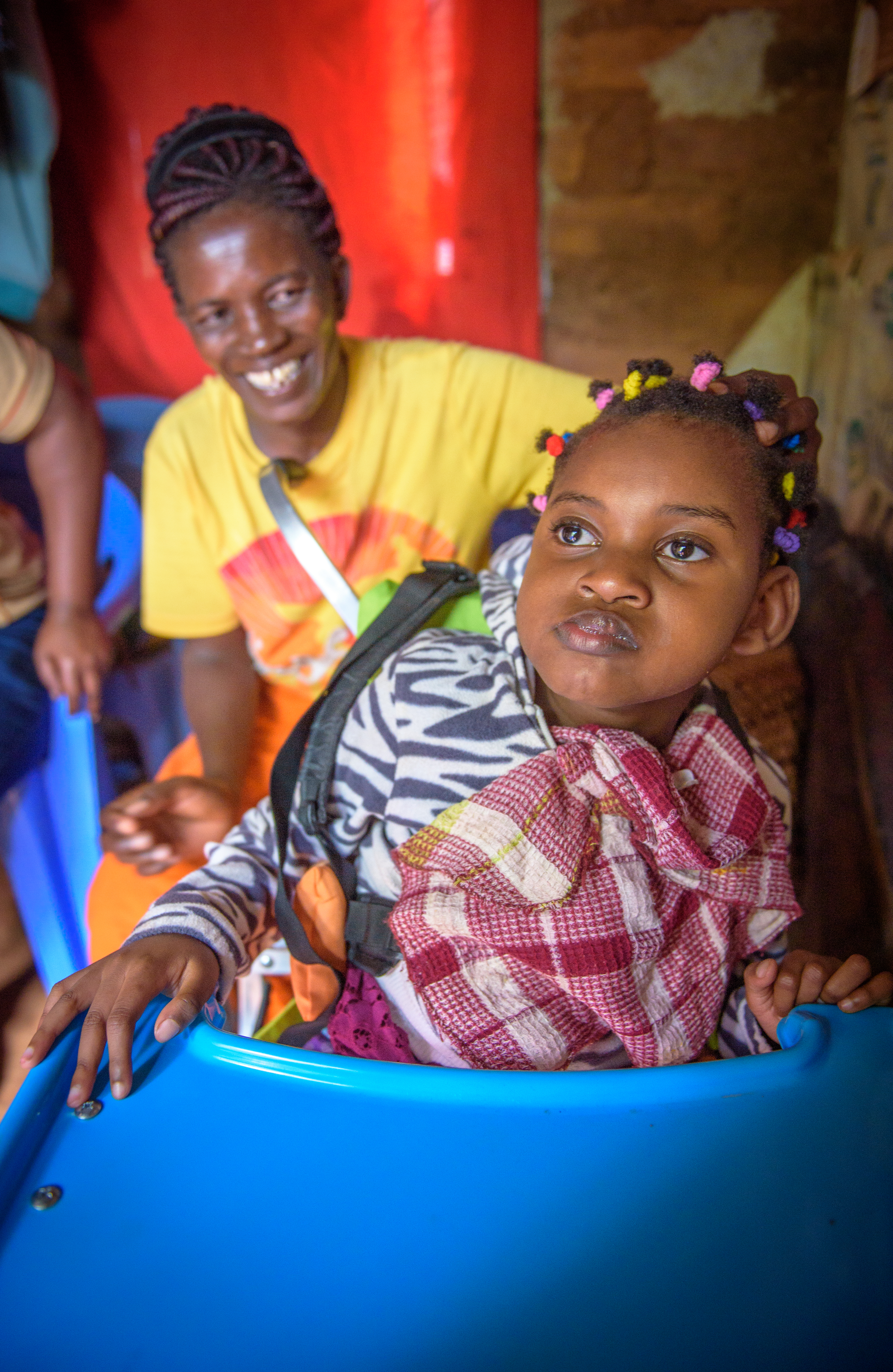 Young Cynthia sits in her wheelchair at home, her mum smiling at her