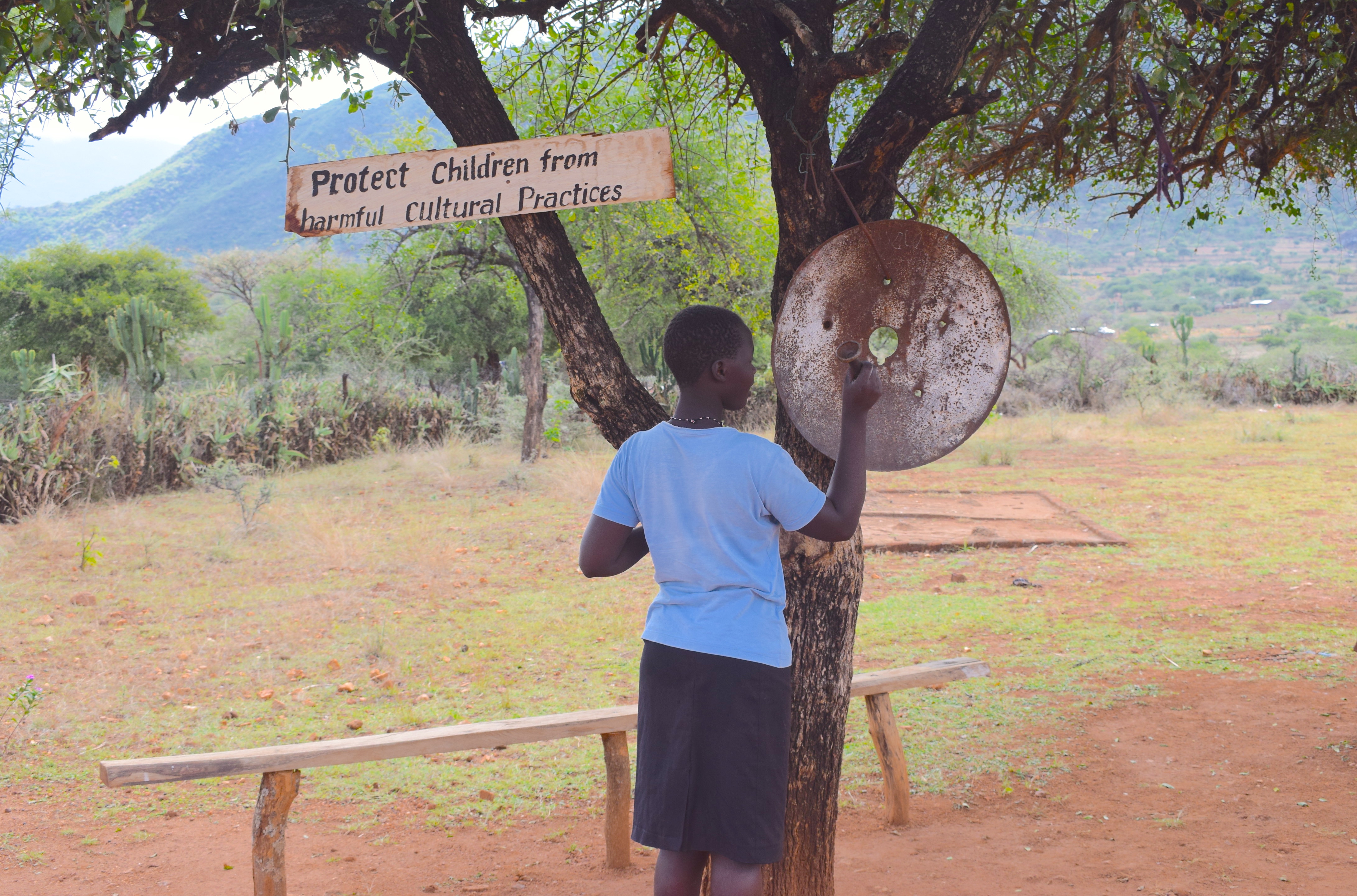A young girl stood under a tree where a sign hangs saying "Protect children from harmful cultural practices.