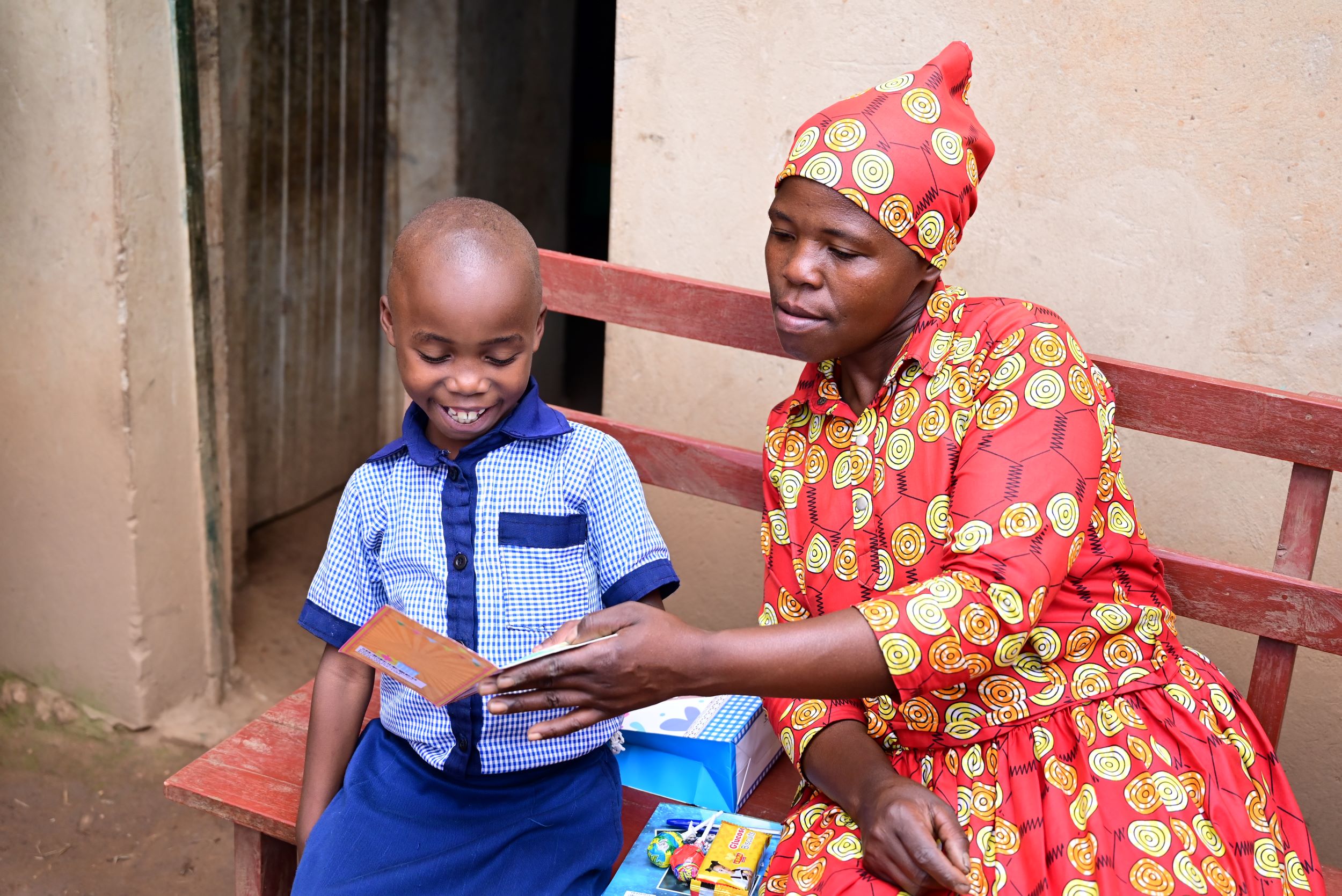 A Rwandan girl in a blue shirt sits on a bench with her mum in a red and yellow dress, reading a message in her birthday card