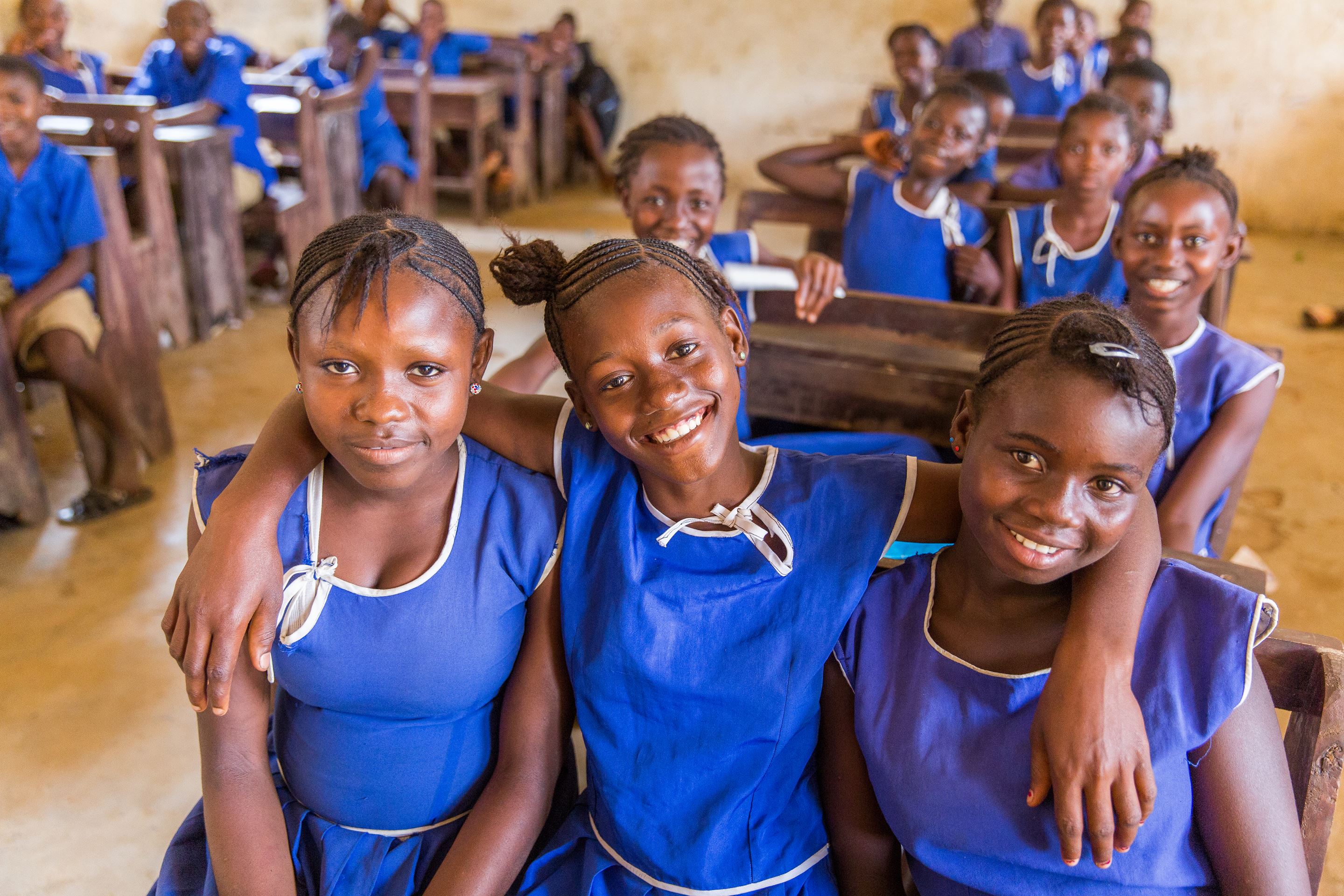 Three children from Sierra Leone smile as they sit in a classroom, arms round each other, wearing blue school tops, with other children in the background