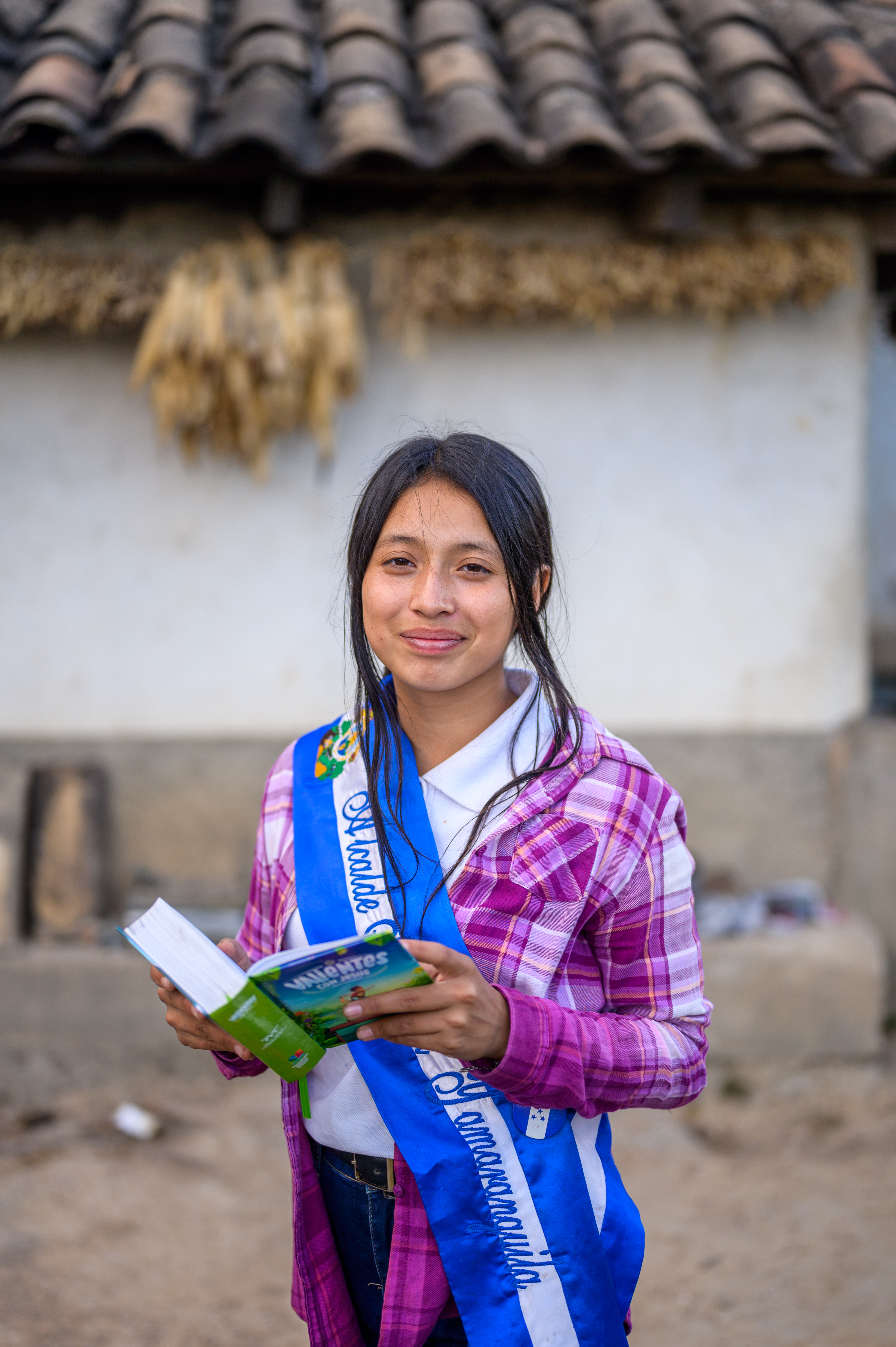 Nahomy, wearing her sash as Youth Mayor, stands in front of her family's small house, with one of her favourite books