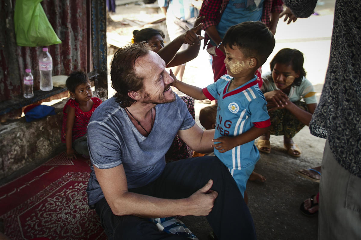 Jerome Flynn sits smiling on a rug on the floor surrounded by children, as a young boy touches his face in Myanmar