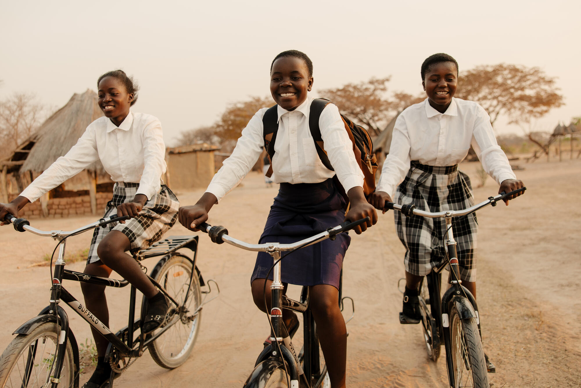 Three girls in school uniforms ride on their bicycles on a dirt track in Zambia; they're smiling to the camera