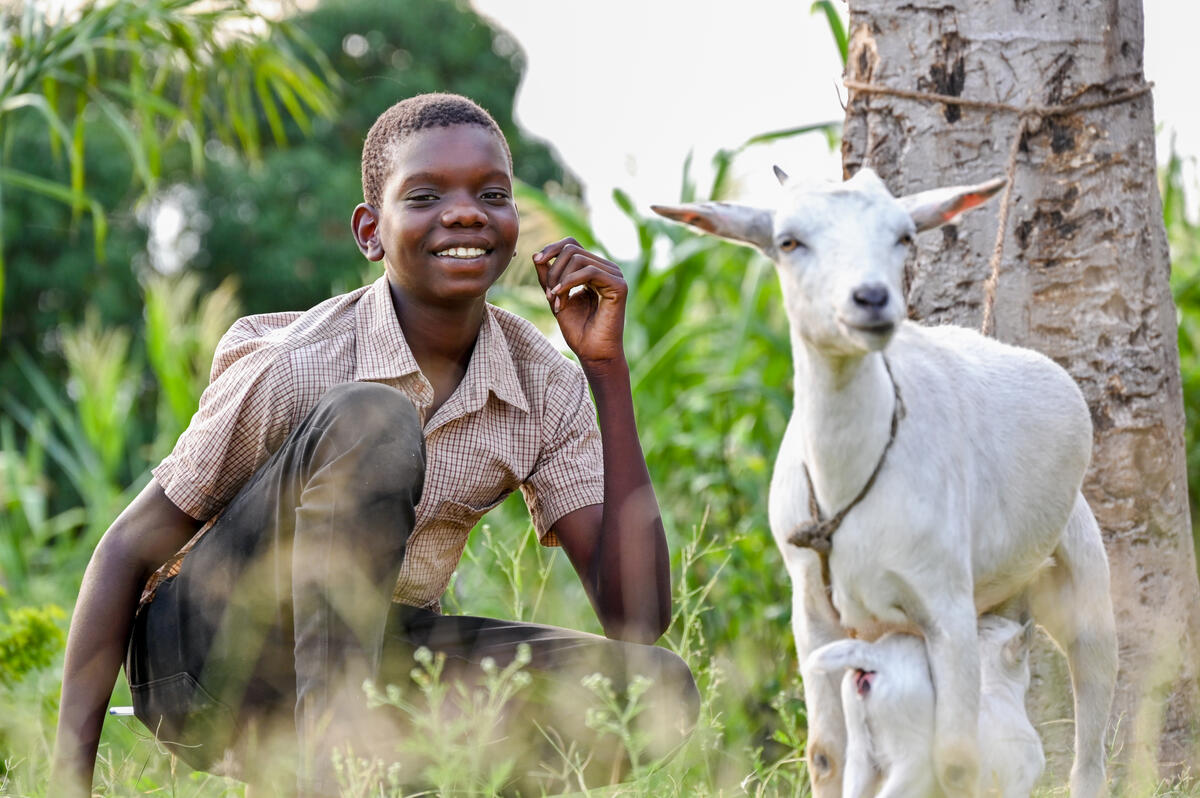 Ugandan teenage boy sat next to a small white goat in a field
