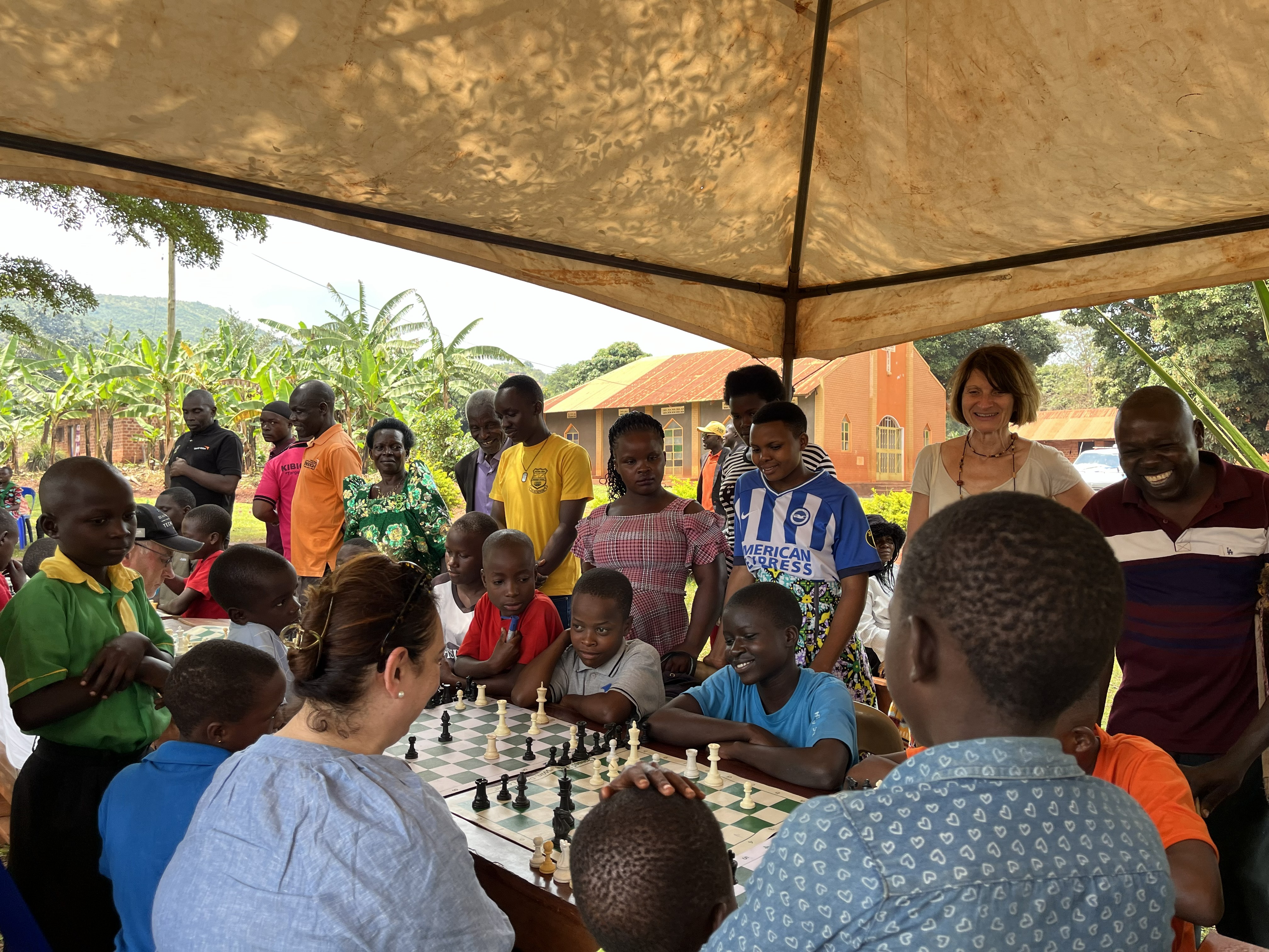 Children play chess at a row of tables while Emmanuelle stands with a group of spectators