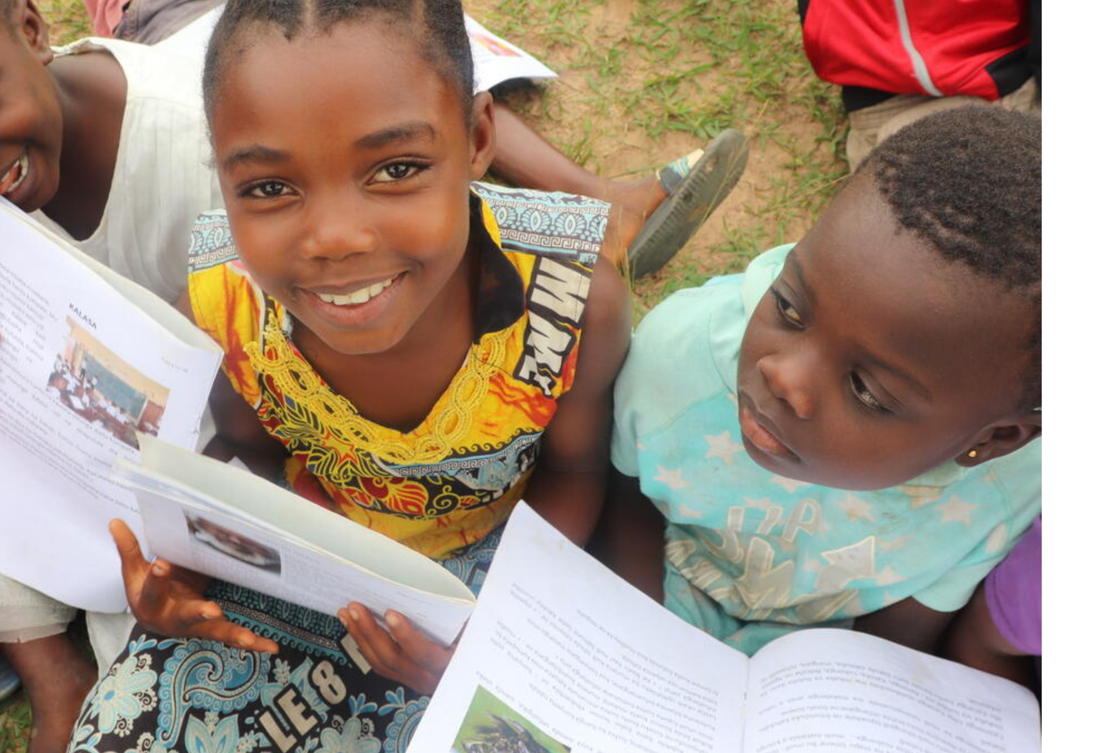 Young girl from DRC sat on the grass, looking up at the camera while holding a book. She is amongst two other children
