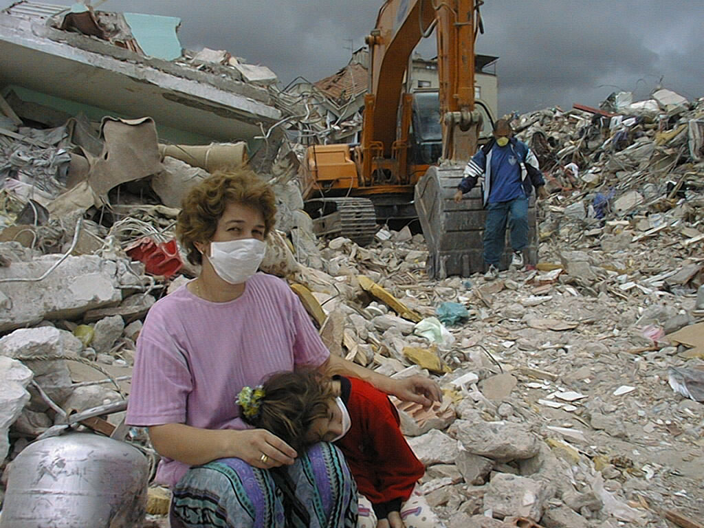 A woman and child sit amidst rubble while a mechanical digger works behind them.