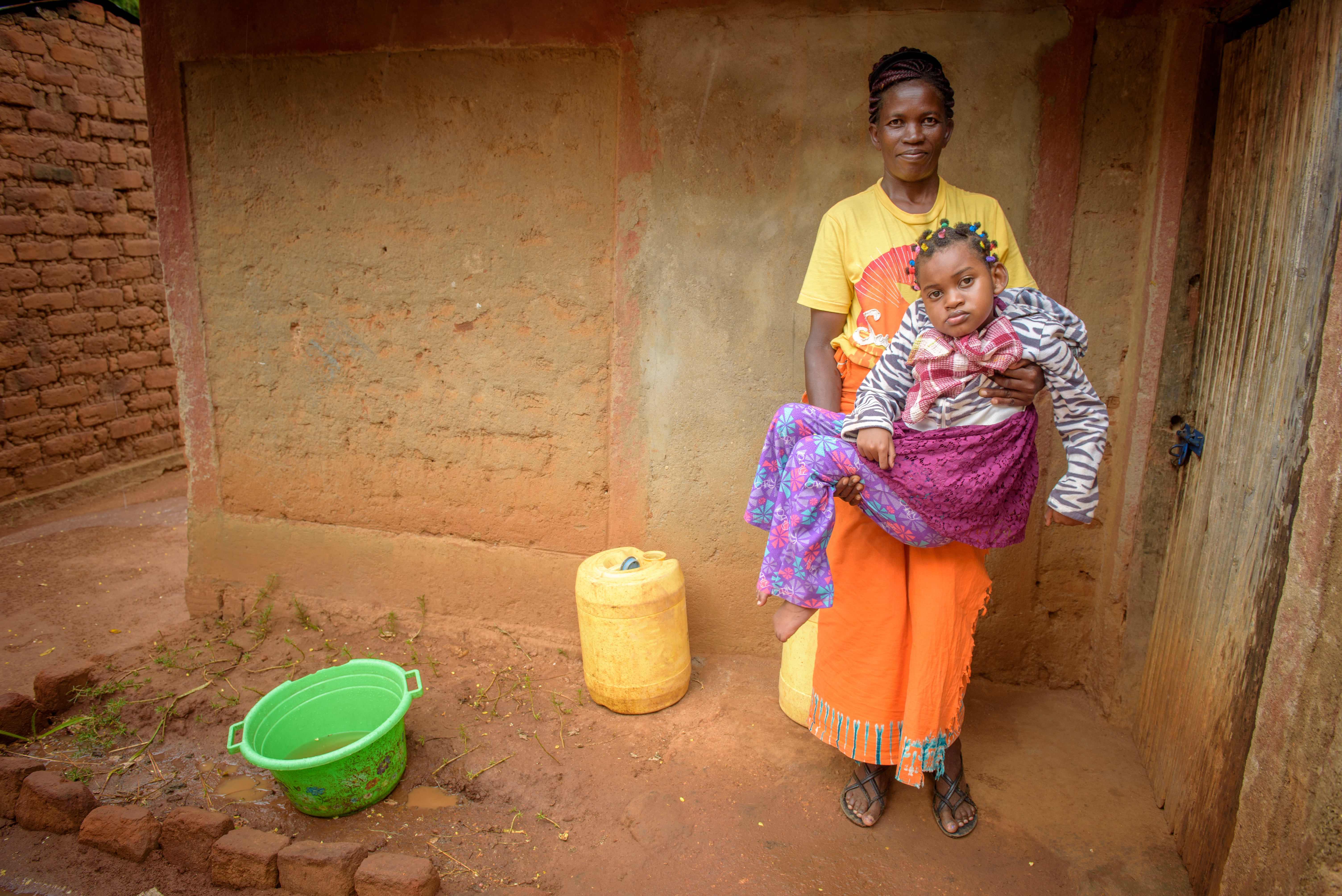 Cynthia being carried by her mum, in their bare dirt home. 