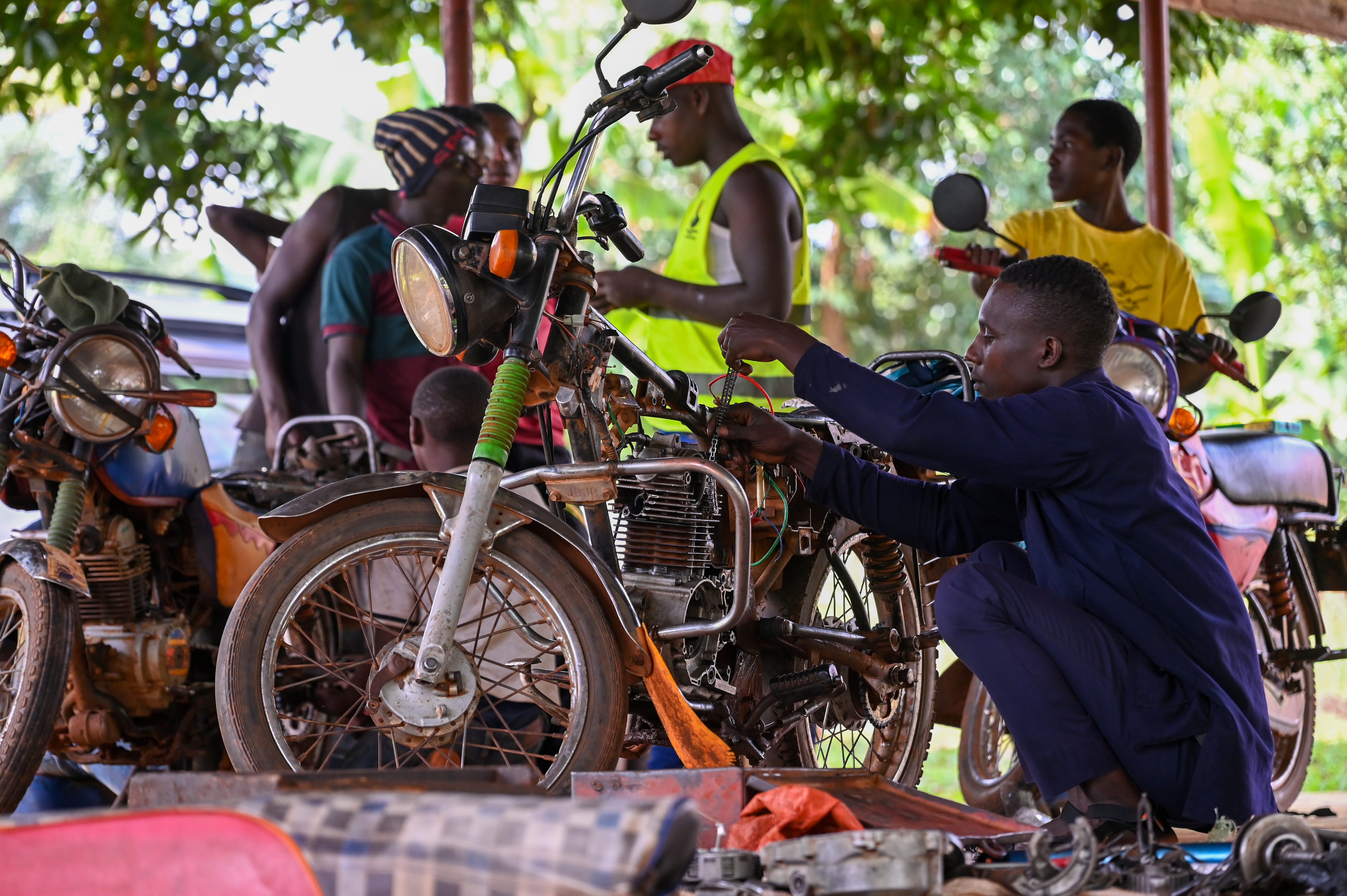 A gentleman in dark blue overalls is crouching, working on a motorbike; a few more men are standing in the background around more motorbikes.