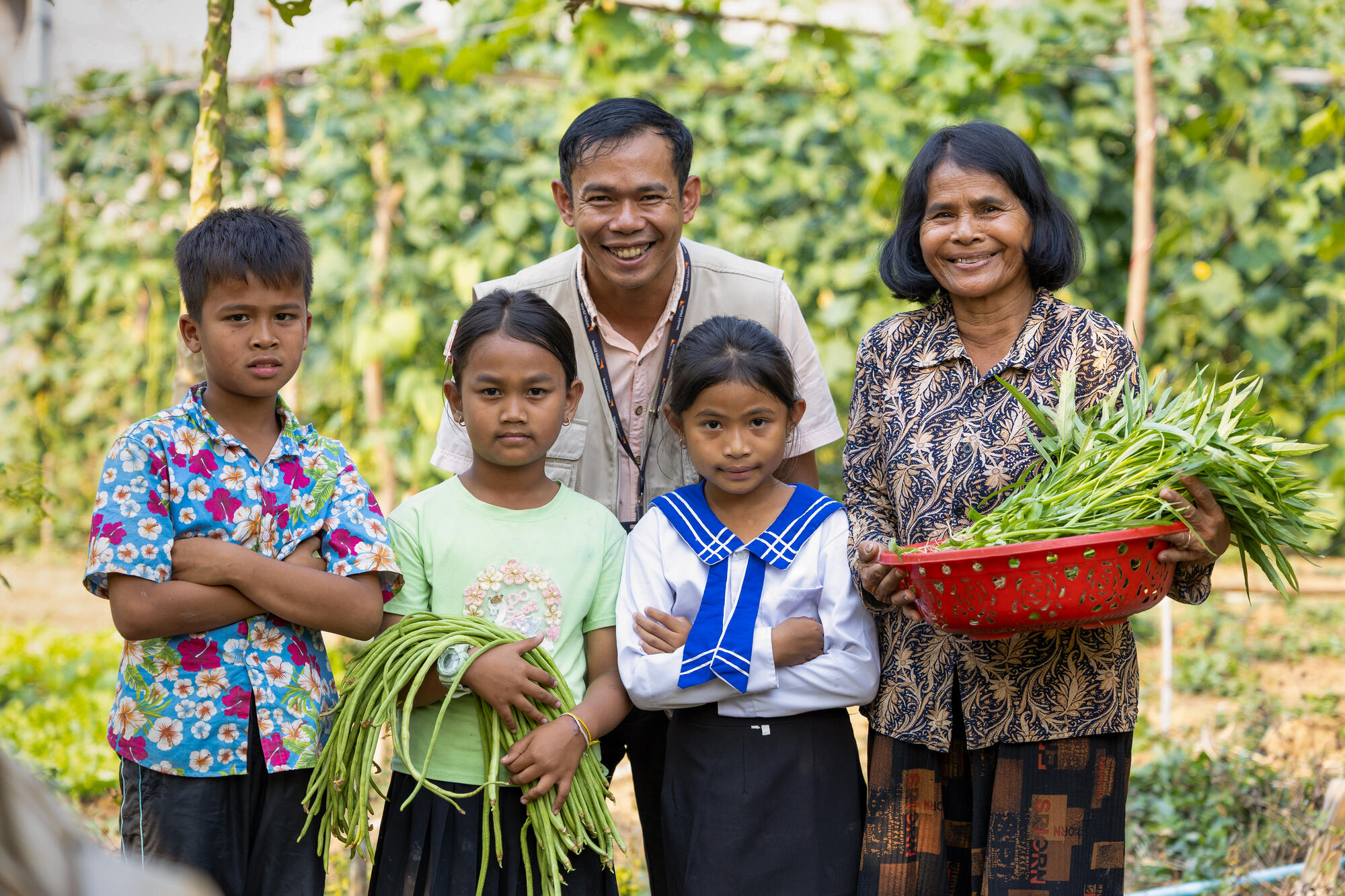 A young girl stands with her grandmother, two friends and a World Vision representative – all facing the camera. They carry beans and other greens they’ve picked in the garden behind them.