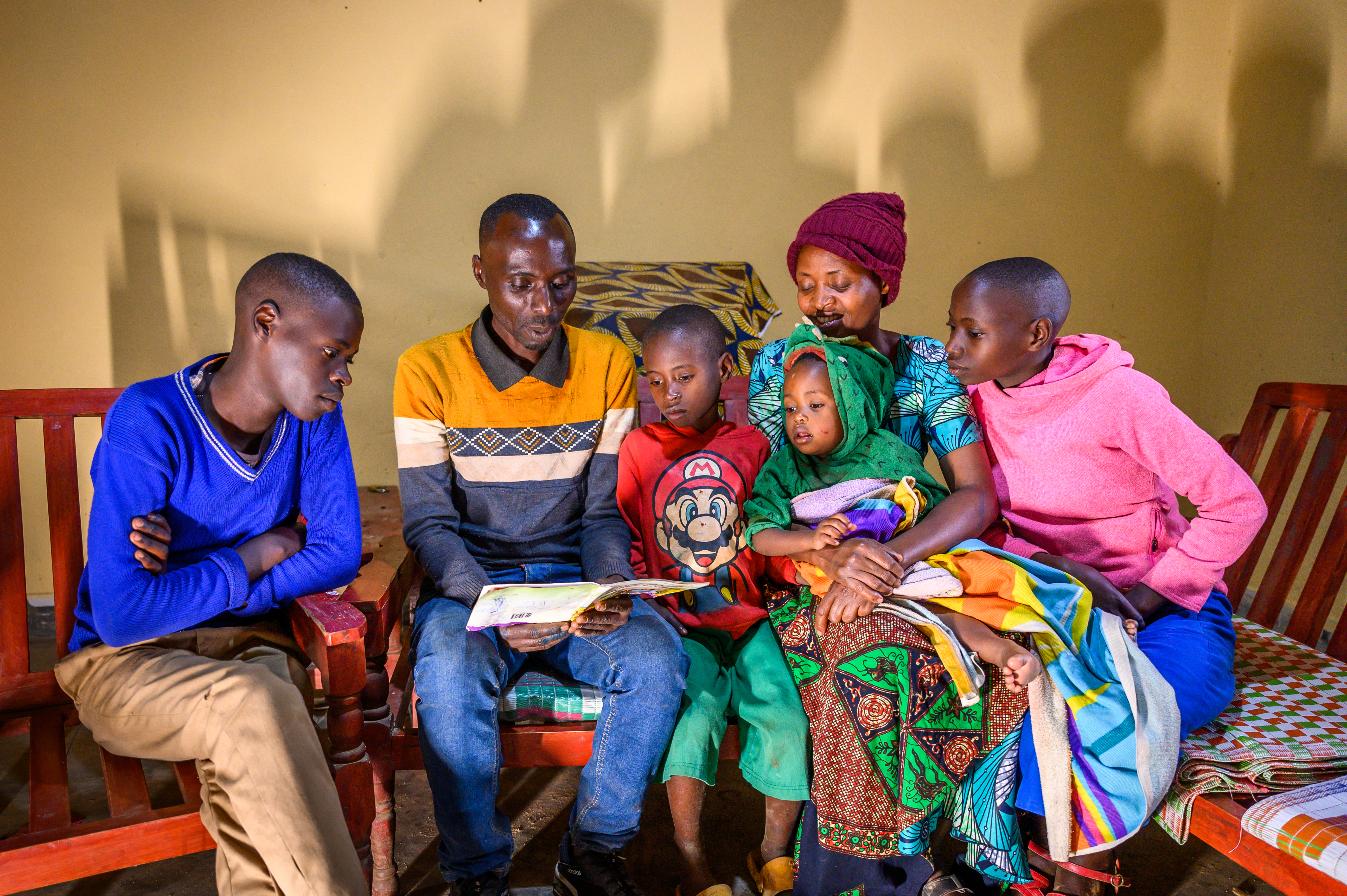 : Juvenal is surrounded by his wife and four of their five children. They’re sitting down as he reads from a book which they all look at with interest.