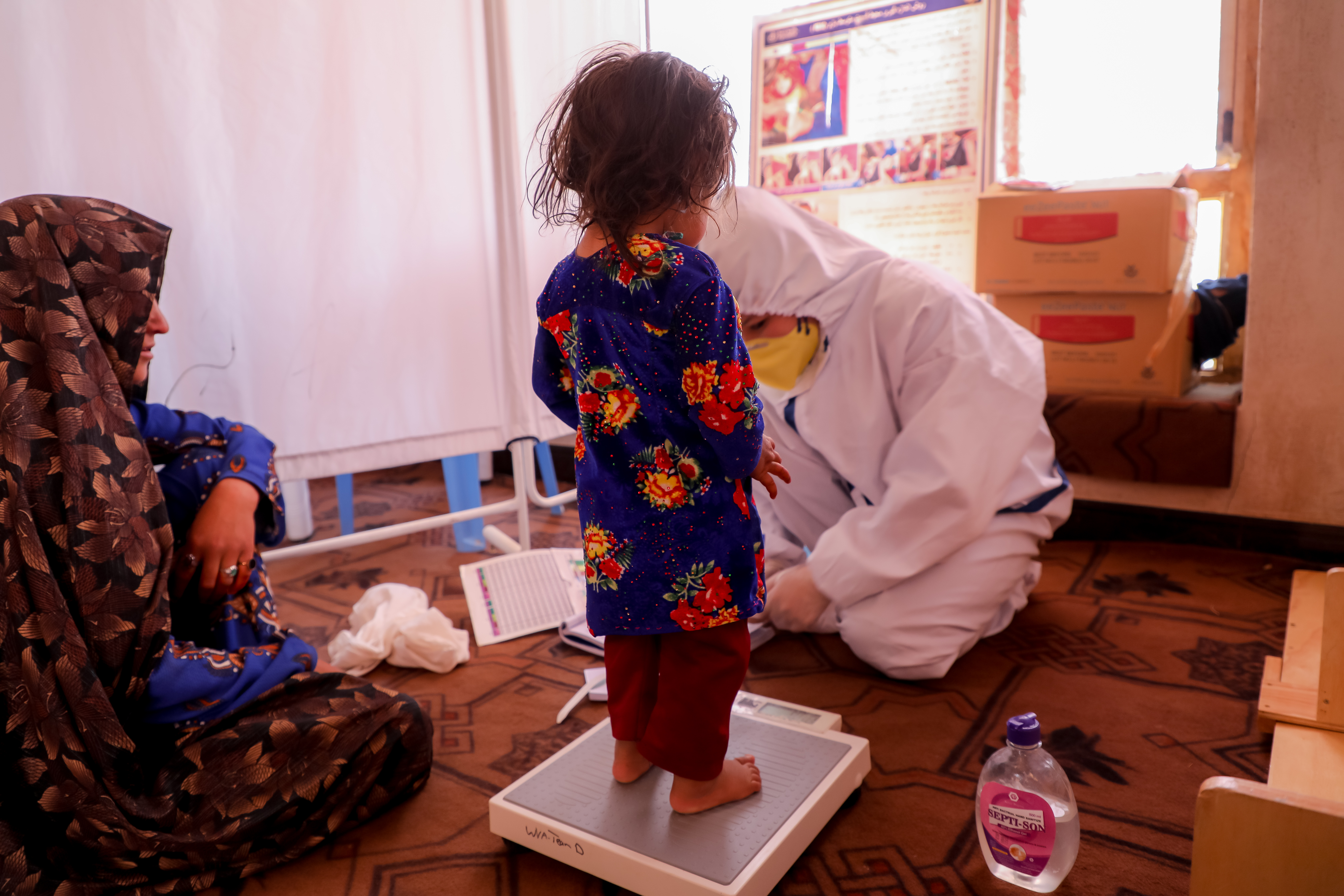 A young child standing on scales being weighed by medical staff, with their mother sat alongside on the floor.