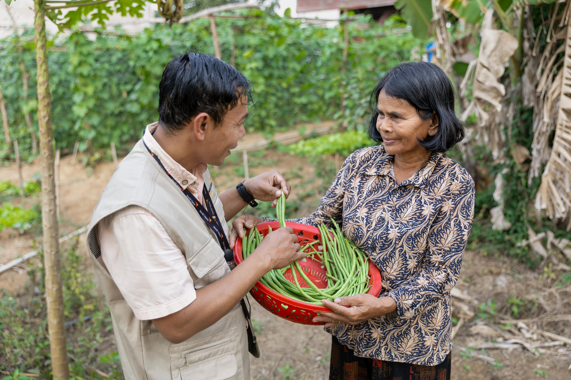 A gentleman is in discussion with a lady. He’s wearing a World Vision lanyard and she’s holding a red plastic basket full of green beans.