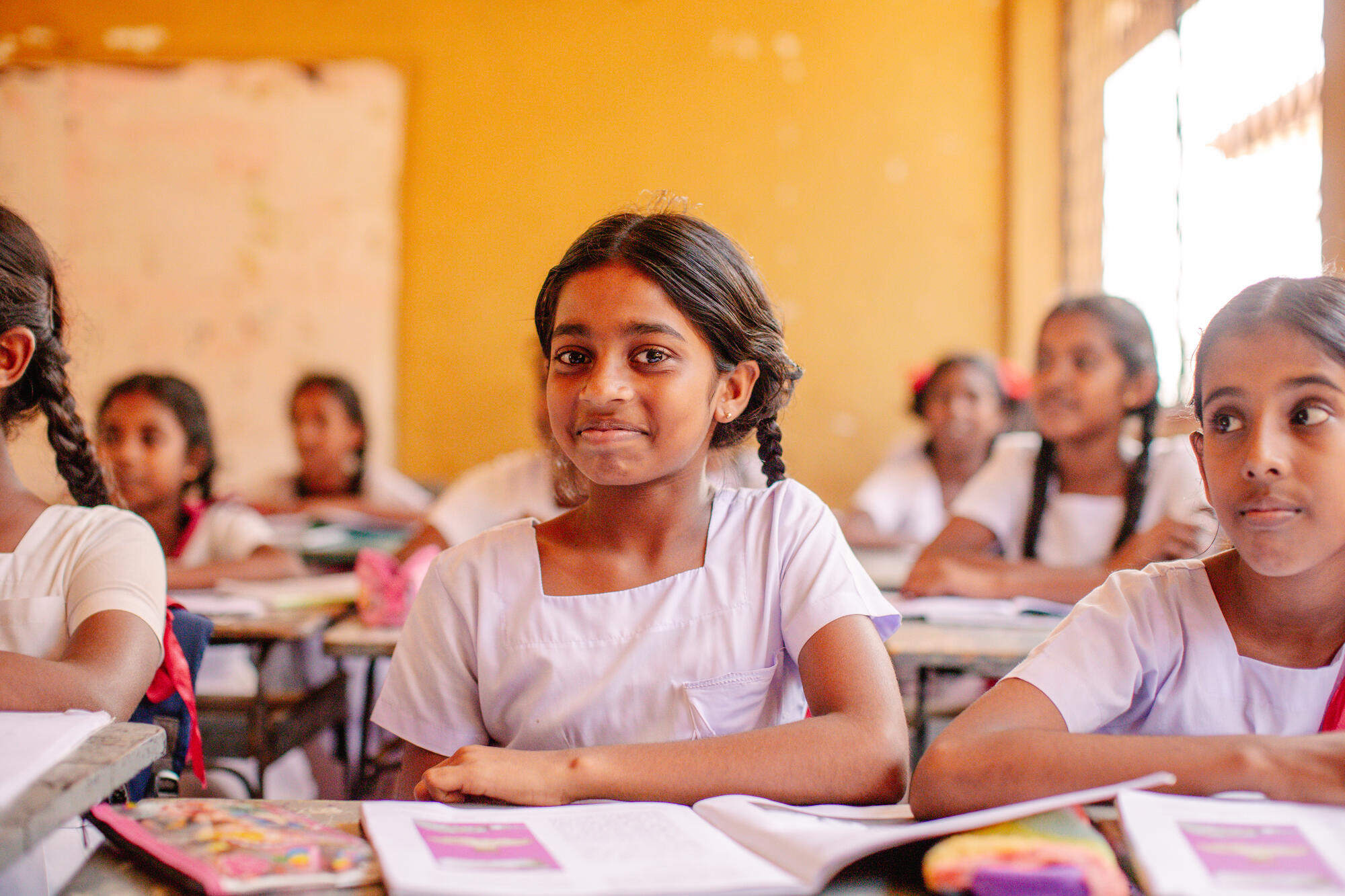 A girl sitting at her school desk with books open smiles to the camera; she's in the middle of a classroom with other pupils listening to the teacher