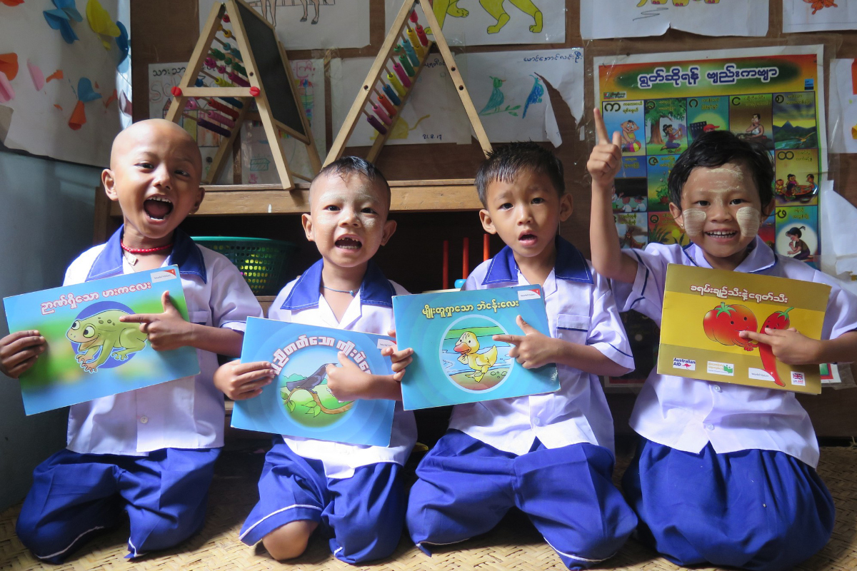 Four children in Myanmar hold books and grin widely as they sit in a classroom
