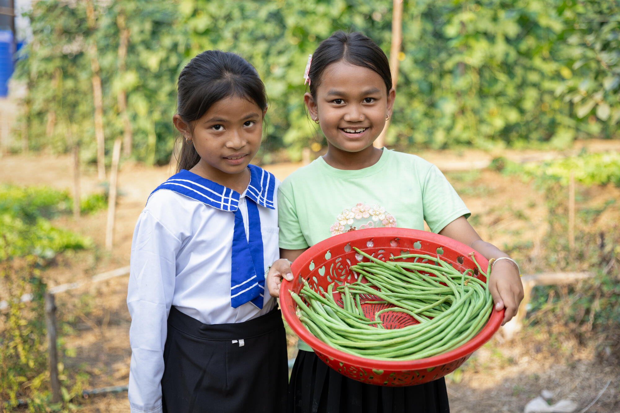 Two young girls are smiling facing the camera, one in her school uniform, the other holding a red plastic basket full of long green beans