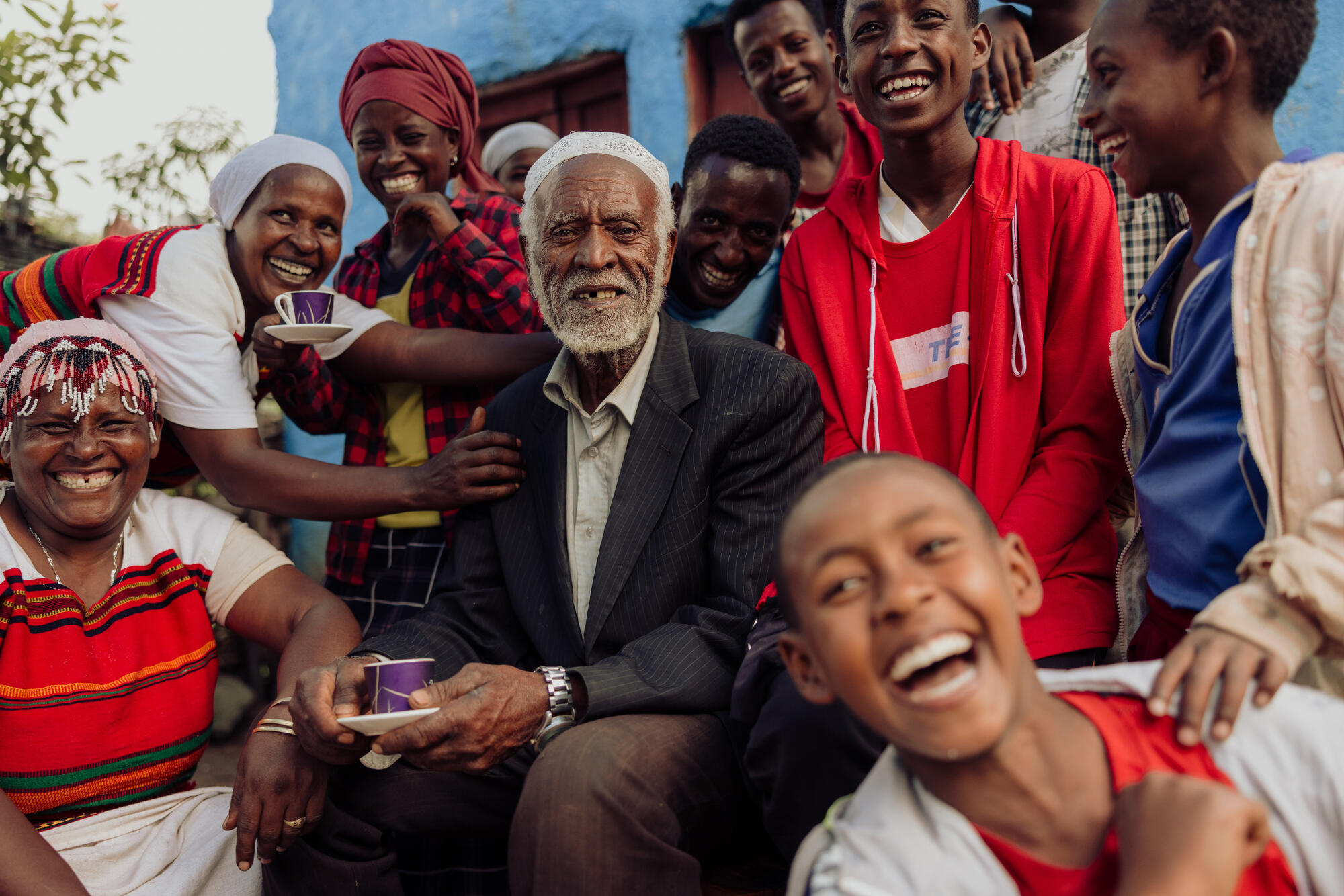 Children and adults surround an elderly gentleman who's sitting with a cup of drink in his hand in Ethiopia: a picture of community togetherness