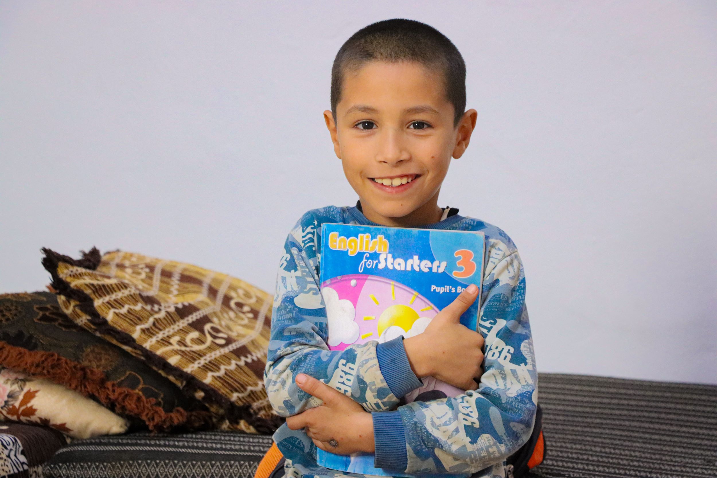 Syrian boy in blue and white jumper holding his book and smiling at camera 