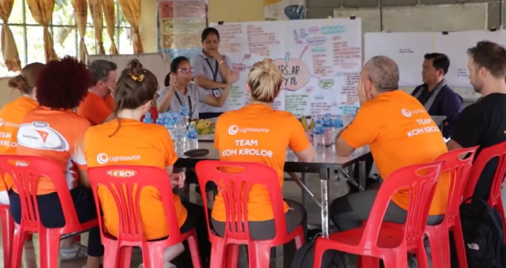 People in orange tops in Cambodia look at a person standing in front of a whiteboard