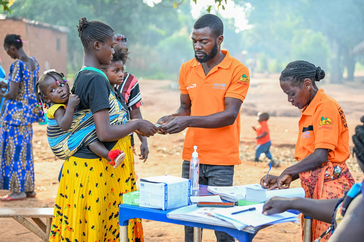 A World Vision health worker hands over medicines to a mother carrying a baby on her back.