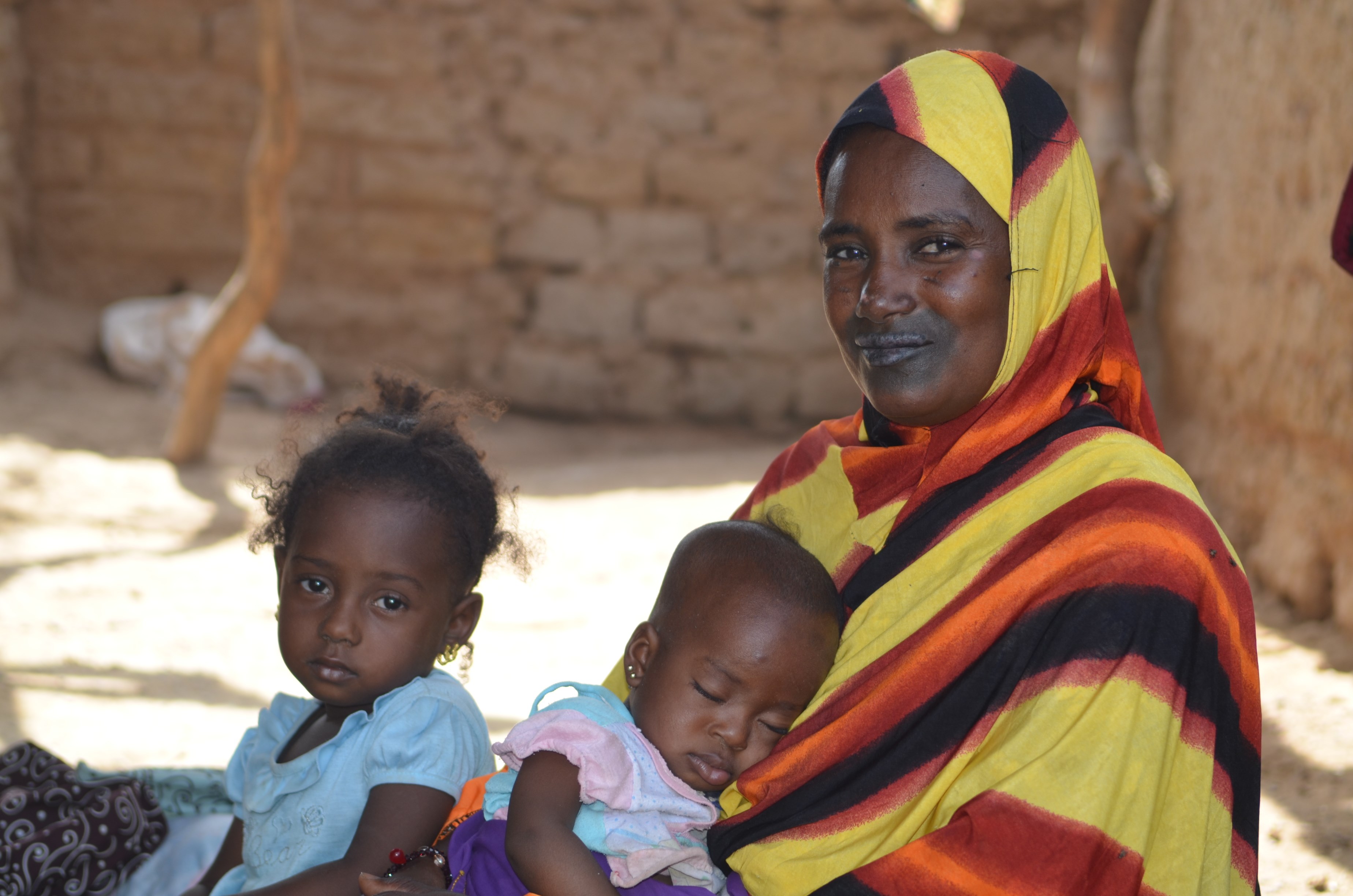 A mother sits in the shade with her two young children