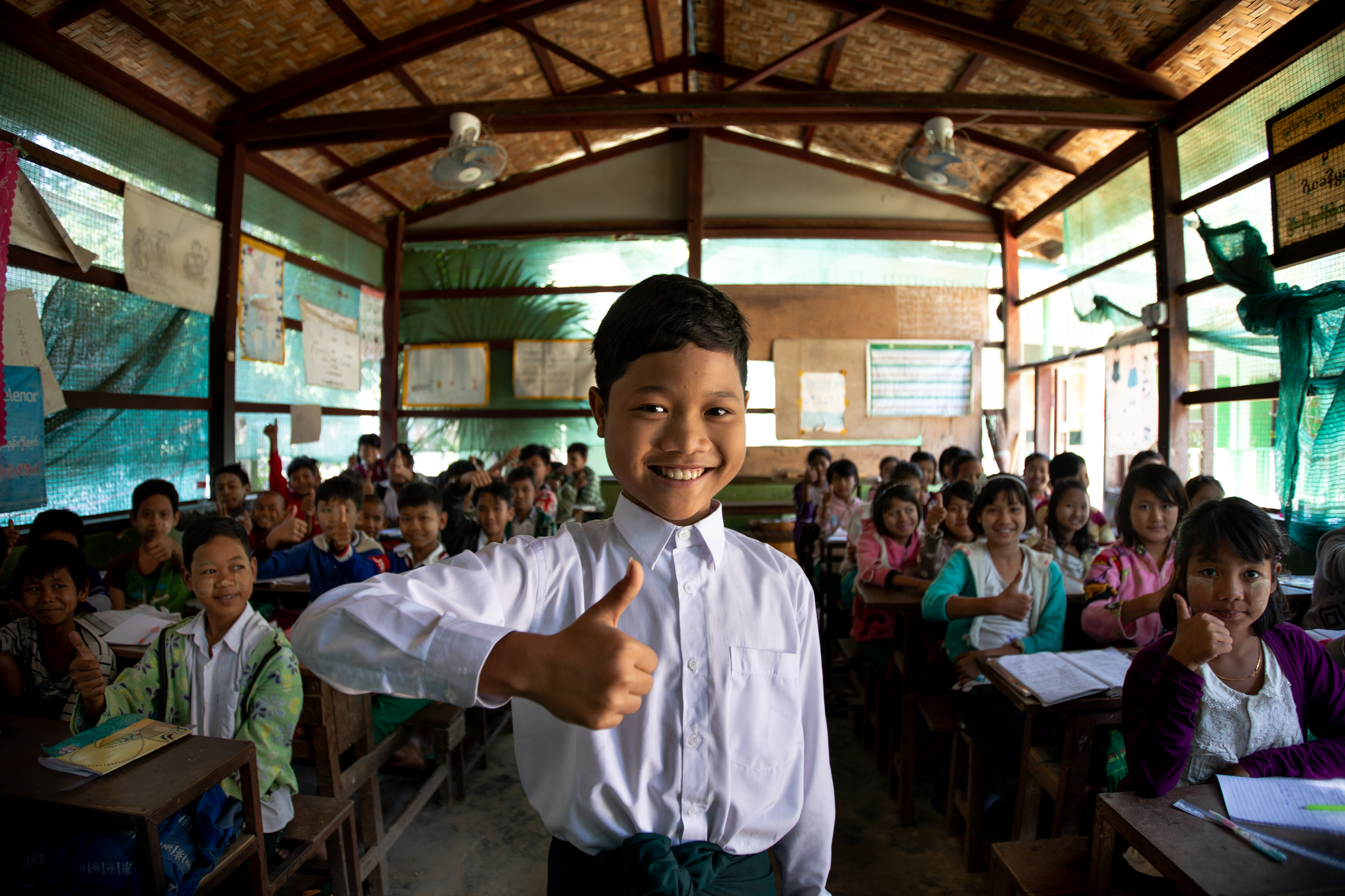 Boy in Myanmar stands at the front of his classroom, giving a thumbs up in front of the rest of his classmates