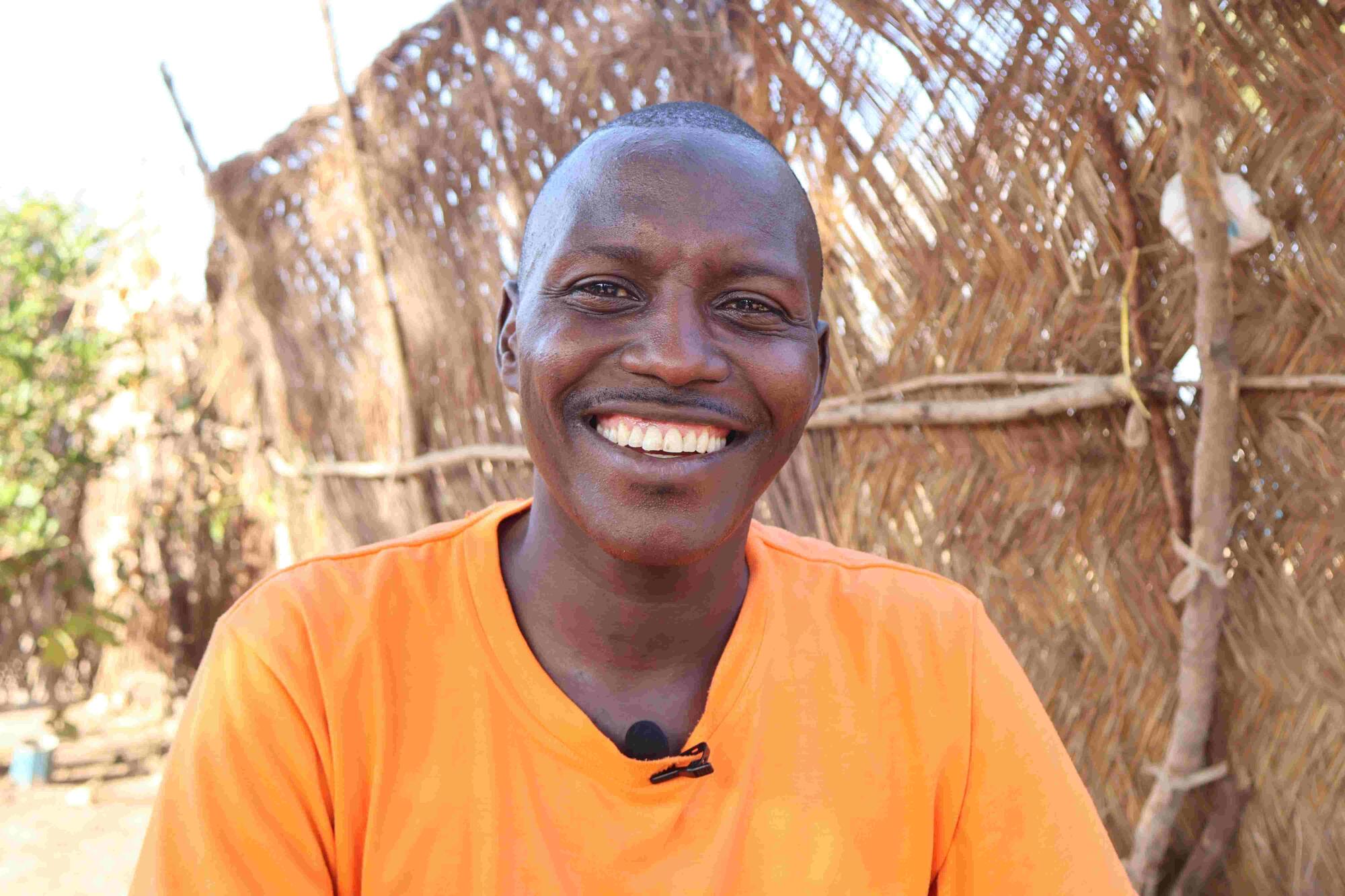 Isa stands outdoors in a refugee settlement in South Sudan: he's wearing an orange tee-shirt and is looking into the camera with a beaming smile
